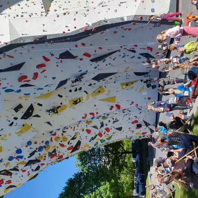 Menschen, die einem Kind beim Klettern einer Wettkampfroute im Sonnenschein zuschauen